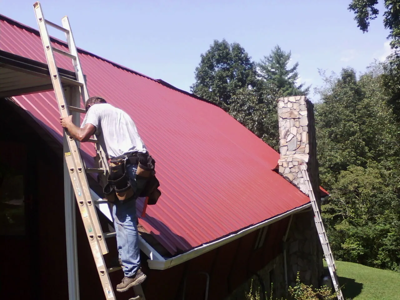 Roofer on a ladder beside a red metal roof and stone chimney — Soffit & Fascia Repair by Roofing Brothers in South New Berlin